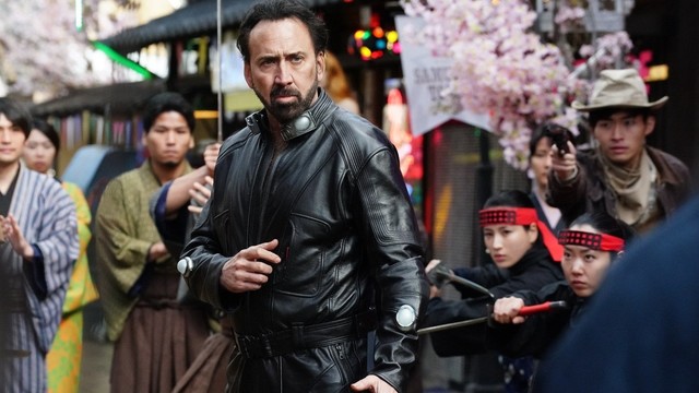 Nicolas Cage in schwarzer Lederjacke steht auf der Straße in mitten mehrerer Japaner, die ihn mit Pistolen und Schwertern angreifen wollen. Er nimmt selbstbewusst eine Kampfpose ein.