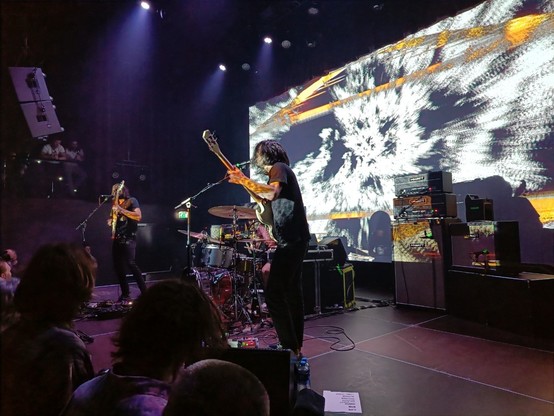 Photo of the band Slift on stage, some people in front of the stage, a big abstract video projection on the screen behind the stage: the video was interactive and responded to the music being played.