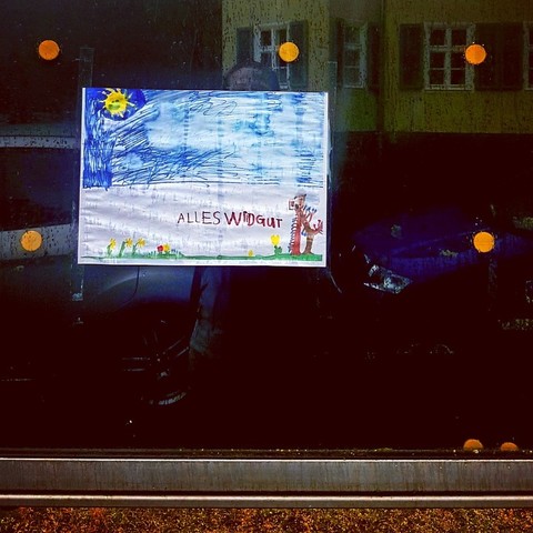 Eine Kinderzeichnung in DIN A4 quer in einer leeren dunklen Info-Schautafel mit vereinzelten Magnet-Buttons. Draußen an einer Straße. Von Feuchtigkeit sind die Farben der Zeichnung bereits verwischt, die einen blauen Himmel mit Sonne zeigt. Die untere weiße Bildhälfte hat einen grünen Streifen mit vereinzelten gelbblühenden Blümchen. Rechts steht ein zeichnerisch juvenil umgesetzter Baum. In der Zeichnung-Mitte von Kinderhand ungelenk gekritzelte Lettern: "Alles wird gut."
