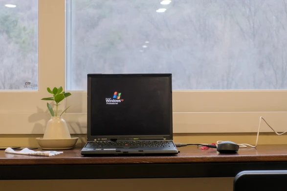 Photo of my Thinkpad X61 standing in front of a window on a wooden counter. On-screen you can see the "Windows XP Professional" logo showing on a black screen, which is one of the basic screensavers Windows XP came with. The laptop is connected to a charger but is currently discharging, something the Lenovo software handles automatically.
