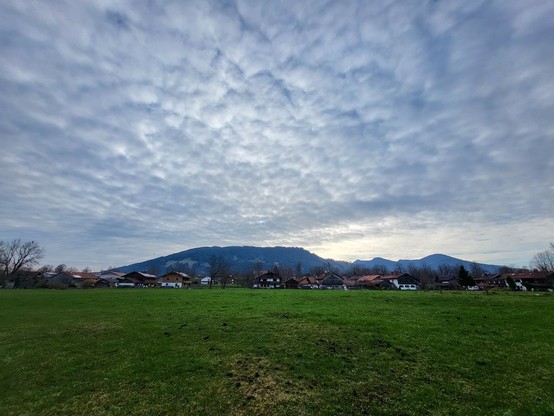 This image captures a serene and picturesque landscape that unfolds across a vibrant green field under a spacious sky, dotted with a few fluffy clouds. The field, lush with grass, stretches into the distance, merging seamlessly with the rural charm of houses nestled within this tranquil setting. In the backdrop, majestic mountains rise, adding a sense of grandeur and permanence to the scene. A variety of trees, including a notable palm tree, punctuate the landscape, contributing to the rich tap…