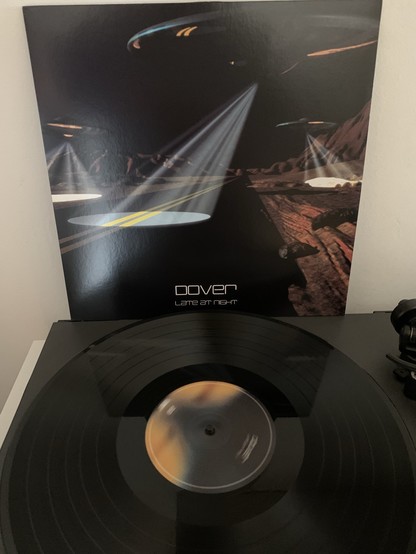 Eine Schallplatte von der Band Dover, das Album  auf einem Plattenspieler, hinter der das Albumcover vom Album „Late at Night“ mit einer Science-Fiction-Szene an der Wand lehnt.