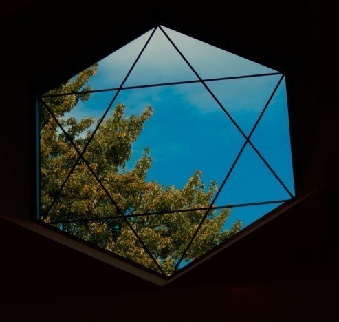 Photo: A hexagonal window with a six-pointed Star of David outlined in it. Outside the window is brilliant blue sky. Running from near the upper left side of the window to the lower right side are the branches of a tree. The branches are covered primarily by green leaves, although some light touches of autumn color — mostly orange — are beginning to show.