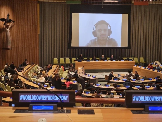This image depicts a significant event held at the United Nations Headquarters in New York, specifically addressing the observance of World Down Syndrome Day. It shows a conference room with attendees seated at desks, focused on a large screen displaying a speaker wearing headphones. The speaker, Puneet, is addressing the gathering on the topic of "Health Equity in the Global South." A prominent feature in the room includes the digital displays on the desks, one of which prominently showcases the hashtag #WORLDDOWNSYNDROMEDAY, emphasizing the event's focus. The setting reflects a formal and collaborative atmosphere, where global issues concerning Down Syndrome and health equity are being discussed and advocated for.