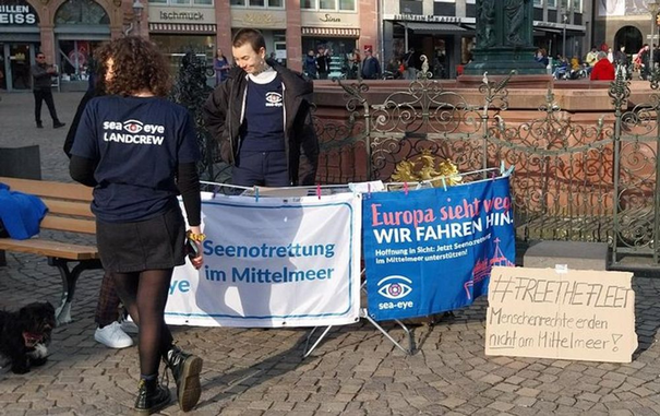 Stand der Sea Eye Landcrew Frankfurt vor dem Gerechtigkeitsbrunnen auf dem Römerberg. Am Stand Transpis mit Sea Eye Logo 'Seenotrettung im Mittelmeer' und 'Europa sieht weg. WIR FAHREN HIN!' Daneben steht ein Pappschild '#FreeTheFleet | Menschenrechte enden NICHT am Mittelmeer!
