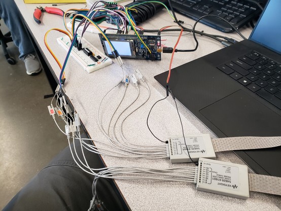 A desk with a laptop, logic analyzer probes, breadboard, and microcontroller development board, connected to an SRAM chip.