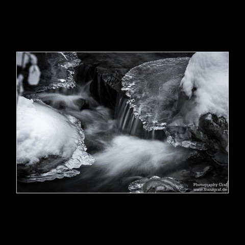 This captivating photograph showcases a serene winter scene focused on a majestic waterfall cascading through a river. The entire landscape is enveloped in a serene blanket of snow, adding to the tranquil and pristine ambiance. The monochrome palette of the image, dominated by shades of black and grey, accentuates the dramatic contrast between the fluid motion of the waterfall and the stillness of the surrounding snow-covered terrain. Notably, amidst this natural beauty, a mammal is subtly pres…