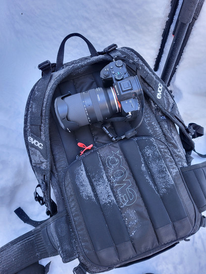 This image captures a serene winter scene where a camera alongside its camera bag rests gently on a blanket of pristine snow. The dominant color scheme of the scene is characterized by the stark contrast between the dark hues of the camera equipment and the camera bag against the pure white snow, emphasizing the solitude and quietness of a winter outdoor setting. The presence of luggage and bags tags suggests that the items are part of a traveler's or photographer's gear, prepared for an advent…