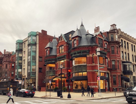 Historic red brick corner building with turrets and a green copper roof, flanked by other traditional buildings, with pedestrians and traffic on the street.