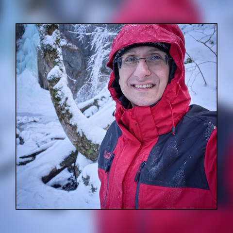 In this vividly captured scene, a man stands amidst a snowy forest, embodying the essence of winter. Cloaked in a striking red coat that contrasts beautifully with the soft pink hues of the snow-blanketed background, he becomes an emblem of warmth in the cold. His face, framed by glasses, carries a gentle smile, suggesting a moment of joy or perhaps a serene appreciation for the surrounding nature. The forest, with its trees lightly dusted in snow, appears as a tranquil backdrop, enhancing the …