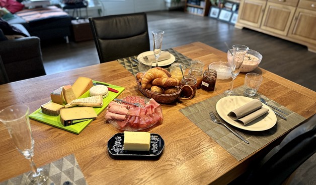Gedeckter Tisch mit Brunch. Eine Platte mit sieben verschiedenen Käsesorten. Kleingebäck, Butter, Konfitüren, Müesli und einem Teller mit verschiedenen Schinken-/Salami-Sorten.
