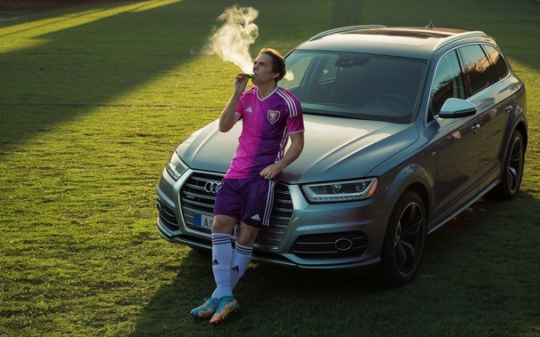 Ein in pink-lila in adidas gekleideter Fußballspieler lehnt an einem dicken Audi SUV und zieht sich einen rein. Alles passiert auf einem Fußballfeld.
