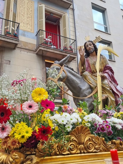 Jesus, auf einem Esel sitzend, wird auf einer mit Blumen und gelben Palmwedeln geschmückten Sänfte durch die Straßen von Barcelona gebuckelt. Dahinter wirbt eine Dame auf einem roten Balkon für ein Erotikmuseum.