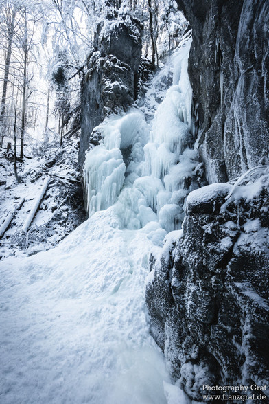 This image captures a breathtaking scene of a frozen waterfall cascading down a rugged cliff. The icy facade of the waterfall glistens against the backdrop of a winter landscape, characterized by snow-covered trees and a blanket of frost that encapsulates the surrounding nature. The atmosphere of the scene is further enhanced by the presence of a blizzard, adding a dynamic and dramatic effect to the winter storm depicted. Amidst this cold and serene setting, the dominant presence of white and b…