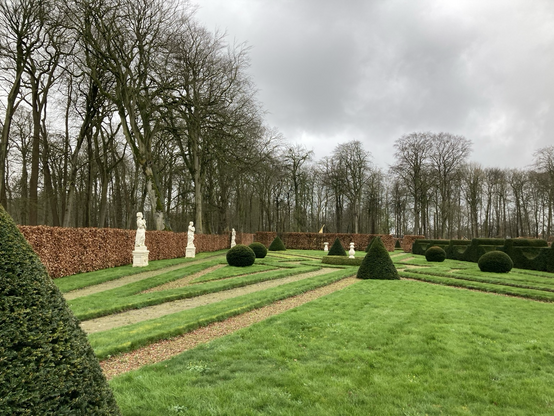 Van links naar rechts: Beukenhaagomheining, daarvoor 3 vd 4 zandstenen vrouwen , taxusbollen, taxuskegels, taxushaag. Achtergrond: hoge bomen vd singel. Onder : gras. boven: grijze hemel 