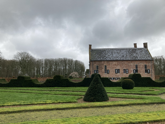 Van onder naar boven: gras met taxusvormen, taxusgolvenhaag, links hoge bomen, rechts achterkant borg met grijs dak, bovenin diverse tinten grijze wolken 