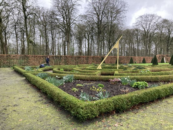 Moderne overgedimensioneerde zonnewijzer in al enigszins door mot aangetaste buxusperken.  Twee hoveniers werken aan de sierbeplanting. Achter hen de haagbeukenhaag, daarachter de wat uit de kluiten gegroeide enorme singelbomen 