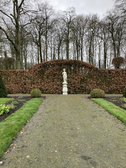 Zandsten beeld van Athene voor mooie boog in de beukenhaag voor de gracht. Zij staat op de dwarsas door huis en tuin. 