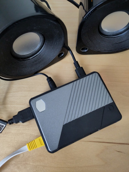 Foto eines Raspberry PI 4-Gehäuses von Cooler Master und zwei kleinen Desktop-Lautsprecherboxen. Der Rechner ist verbunden mit einem USB-C-, Klinken-, USB-A- und LAN-Kabel.