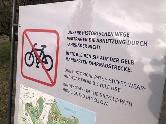 Plakataufsteller an einem Parkeingang in Potsdam, Hinweis mit durchgestrichenem Fahrrad-Symbol: 
„Unsere historischen Wege vertragen die Abnutzung durch Fahrräder nicht.“ Un d dann noch „Bitte bleiben Sie auf der gelb markierten Fahrradstrecke”.