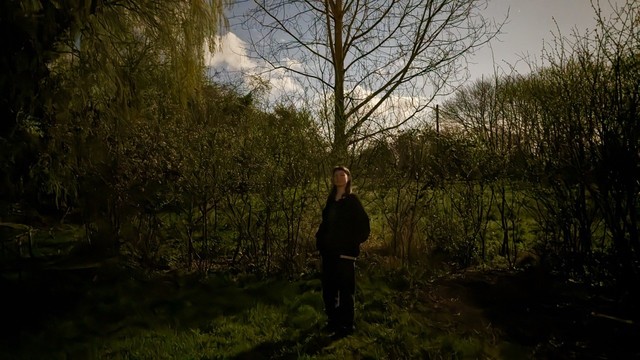 A woman stands in a garden in the night-time. There is with very thin bare early foliage on the trees and hedge behind her. The sky looks blue but it is dark. 