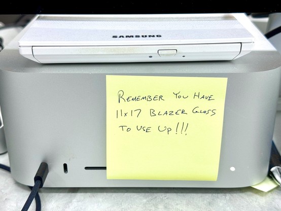 A yellow Post-it® brand sticky note is attached to an M1 Mac Studio computer. The message written on the note is, “Remember you have 11×17 Blazer Gloss to use up!!!”

Unrelated: a white Samsung tray-loading Blu-ray disc reader/writer is on top of the Mac Studio.