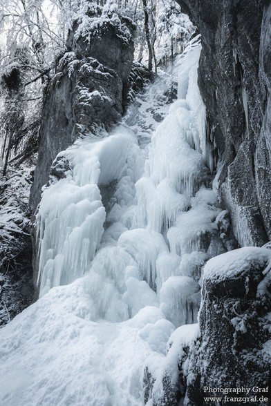 This captivating image showcases the serene beauty of a frozen waterfall nestled amidst a winter landscape. The scene is enveloped in a blanket of snow, emphasizing the season's quiet and peaceful ambiance. Dominated by shades of grey and white, the photograph presents a harmonious blend of the winter's chill and nature's grace. The waterfall, now a sculpture of ice, becomes the central figure, surrounded by snow-covered trees that stand as silent guardians of this secluded spot. The stream tha…