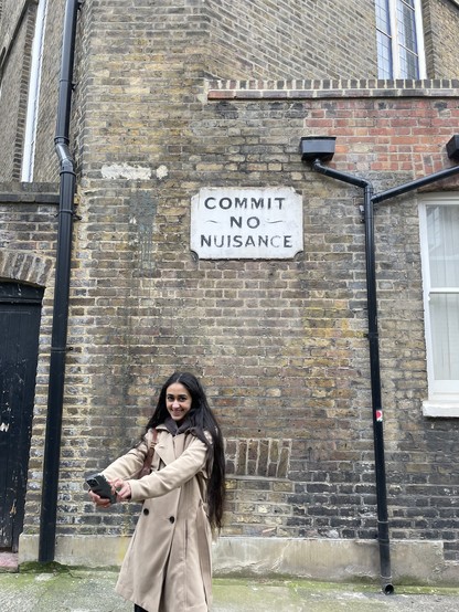 Woman holding her phone in front of her with a brick building behind her. On the wall of the building is a white sign with scalloped corners and simple black block letters that read "commite no nuisance".