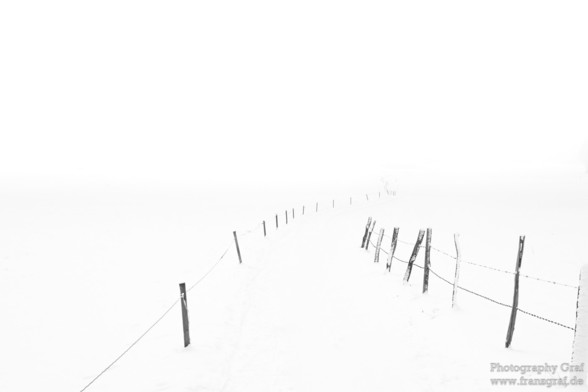 This image captures a serene, winter scene dominated by the simplicity and quiet beauty of a fence blanketed in snow. The atmosphere is enveloped in fog, adding a layer of tranquility and mystery to the landscape. The entire composition is rendered in black and white, emphasizing the stark contrast between the snow-covered ground and the dark lines of the fence, while also highlighting the subtle textures of the winter landscape. The absence of color enhances the monochromatic beauty, drawing a…
