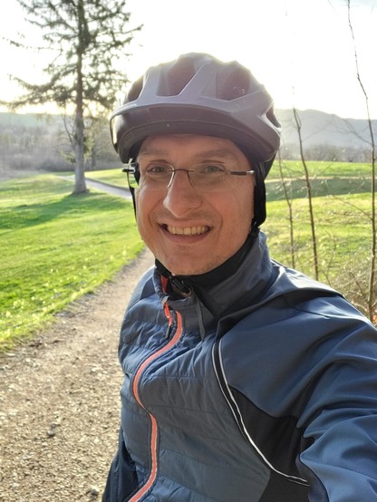 In this vibrant outdoor setting, a man exudes a sense of adventure and joy, capturing a selfie moment that feels both personal and expansive. Dressed for an active day out, he's clad in attire suitable for hiking, including a helmet that speaks to his readiness for any physical challenges the day might present. His smile, genuine and wide, reflects the happiness of being immersed in nature. Surrounding him, the lush greenery of grass and trees paints a picture of a day well-spent outdoors, unde…