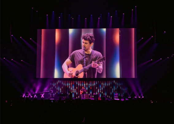 Image of John Mayer on stage with projected image behind on a giant screen and lights surrounding it. 