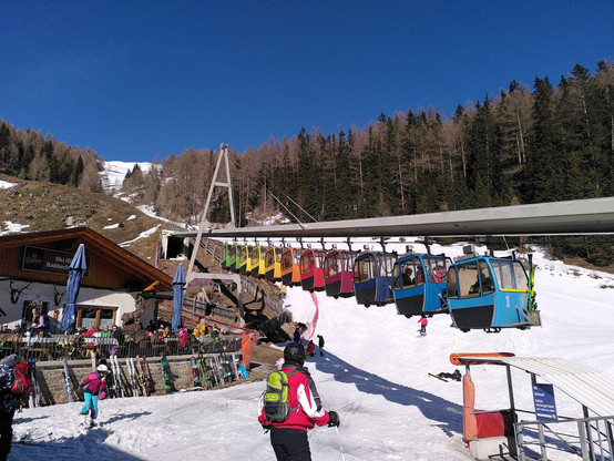 Aus 11 einzelnen Kabinen, jede ein einer anderen Farbe, vorn grün über gelb und rot bis blau, bestehende Hängestandseilbahn ist soeben aus einem Tunnelportal im Hang gekommen und quert eine daraunter liegende Skipiste 