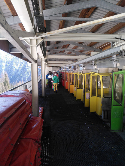 Talstation der Hängestandseilbahn von innen mit in der Station stehender Garnitur, bestehend aus 11 Einzelkabinen, die Türen sind offen, wintersportausübende Personen verlassen den aus Matten bestehenden, ebenerdigen und nicht erhöhten "Bahnsteig" nach hinten