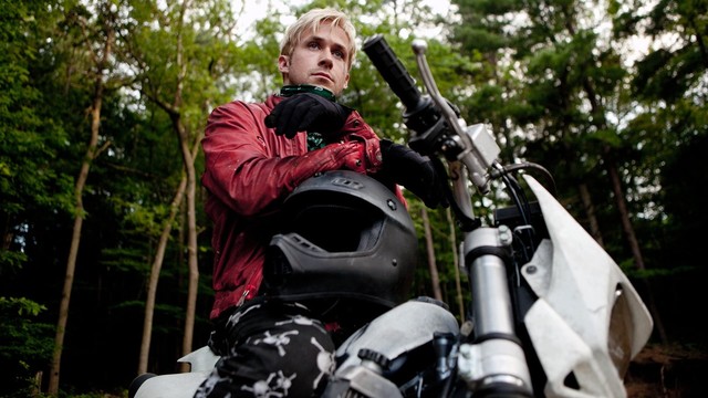 Ryan Gosling mit wasserstoffblonden Haaren sitzt mit roter Lederjacke auf einem weißen Cross-Motorrad, auf dem Tank für sich ein schwarzen Vollintegralhelm, im Hintergrund Wald. Die Kamera blickt fast von Bodenhöhe zu ihm hoch.