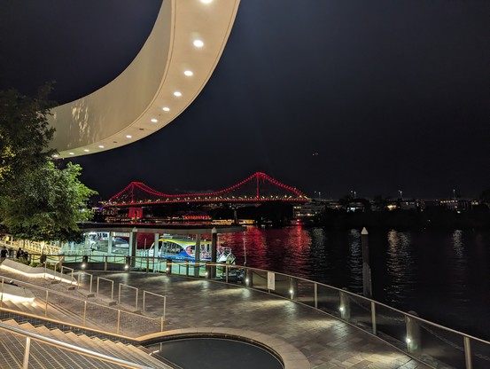 Story bridge over the Brisbane River kit up in red and white.
Decorative curved off white flying gantry in the foreground with downlights picking out steps down to the bikeway asking the river. 
Lights from the far side reject in the river.