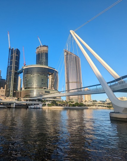 Looking north across the Brisbane River. 
In the foreground, a pedestrian suspension bridge under construction. 
In the background, glass towers under construction in the CBD.
Tall cranes against a clear blue sky.