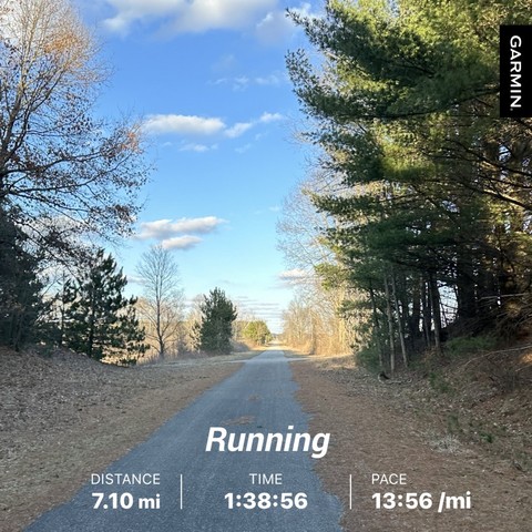 A paved trail with trees on either side, under a blue sky with clouds. A Garmin fitness tracker overlay shows a running distance of 7.10 miles, time of 1:38:56, and pace of 13:56 per mile
