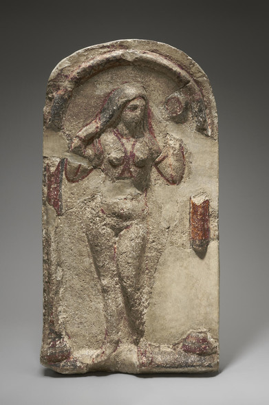 Limestone relief of Aphrodite in a niche, Roman artwork of 200-256 CE. The goddess is standing in the nude, wringing out her hair as if emerging from the bath or the sea. Red lines of paint suggest jewellery like a necklace and a body chain between the breasts.