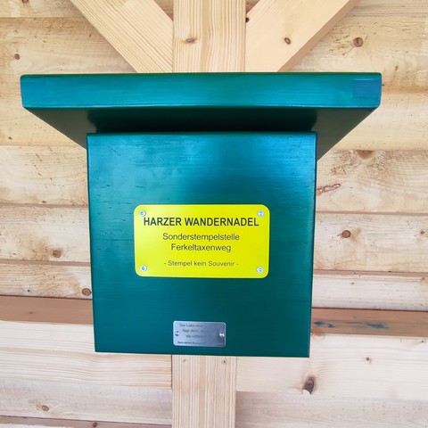 In einer holzhütte hängt ein grüner holzkasten mit einem gelben Schild

Aufschrift:
HARZER WANDERNADEL
SonderStempelstelle Ferkeltaxenradweg
Stempel kein Souvenir