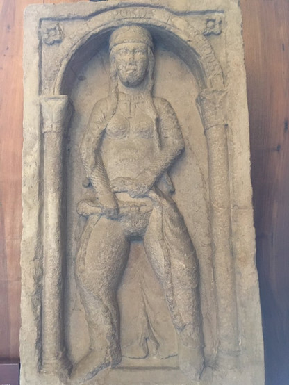 Relief of a woman, fully dressed, who is lifting her skirt to, by the looks of it, shave off her pubic hair. She is a respectable woman wearing a headdress.