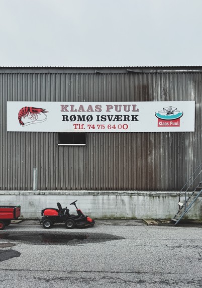 DE: Fabrikhalle eines Krabbenfischers im Hafen von Havneby auf Rømø, Dänemark. Auf dem Firmenschild steht „Klaas Puul, Rømø Isværk“ und eine Telefonnummer. Vor der Halle ist ein schwarz-roter Rasenmähtraktor mitsamt rotem Anhänger abgestellt, am rechten Bildrand sind Stufen einer Eisentreppe zu sehen.

EN: Factory building of a crab fisherman in the harbour of Havneby on Rømø, Denmark. The company sign reads "Klaas Puul, Rømø Isværk" and a telephone number. A black and red lawnmower tractor with a red trailer is parked in front of the hall and the steps of an iron staircase can be seen on the right-hand side of the picture.

