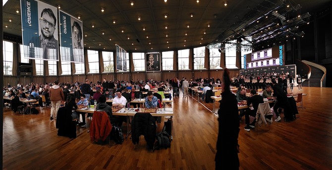 Main event hall with players seated at hundreds of tables playing chess, incl a stage where the top 9 games are being played.