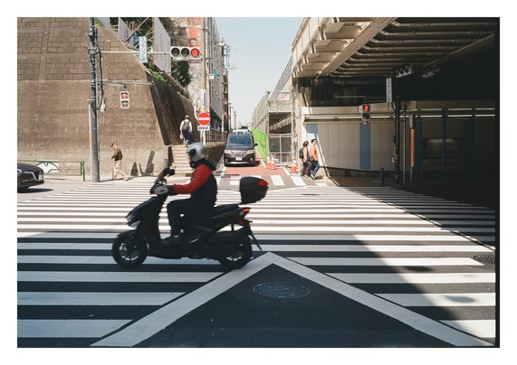 A motorscooter with a red jacket speeding across a pedestrian crossing