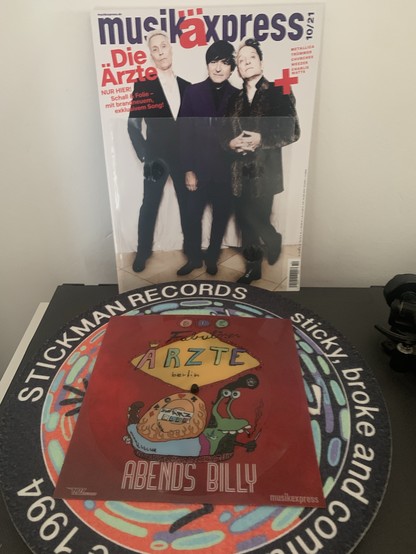 Ein Foto von der Flexidisc von „Abends Billy“ auf einem Plattenteller, im Hintergrund die Ausgabe der Zeitschrift „Musikexpresse“, in der diese erschienen ist, mit der Band „Die Ärzte“ auf dem Cover.