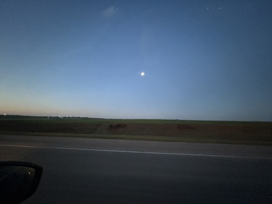 Dusk view from a moving vehicle showing a flat horizon, a moon in a darkening sky, and distant lights. A section of the vehicle's window frame is visible at the bottom left.