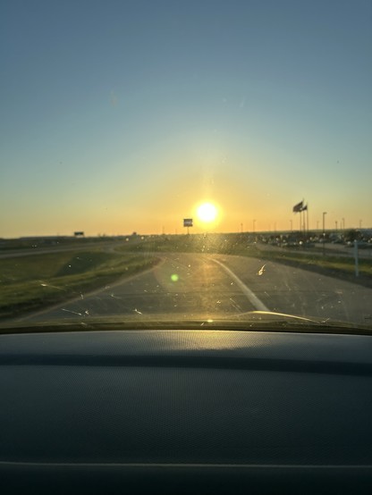 Sunrise view from inside a car with sun visor visible, overlooking a road leading towards the sun with flags in the background.