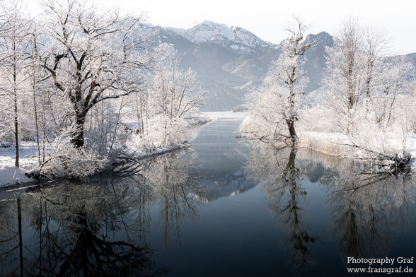 This captivating image illustrates a serene river gracefully meandering through a stunning landscape adorned with trees. The scene is further embellished by the majestic mountains in the background, which are cloaked in a pristine layer of snow, reflecting the quintessence of winter's beauty. The trees, dusted with snow, stand tall, adding a touch of verdant green amidst the predominantly white and grey hues of the cold season. The sky above is clear, hinting at the crisp freshness of the air, …