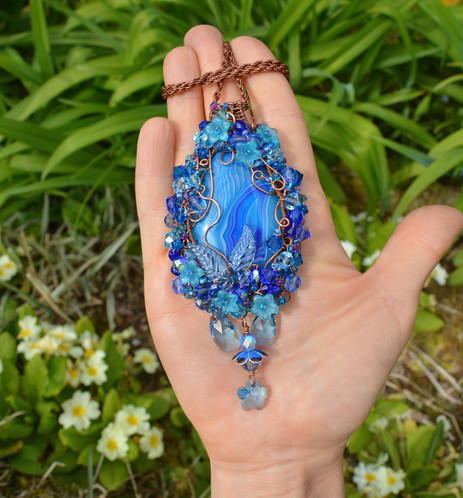 An elaborate blue pendant necklace, resting on an open hand. The pendant is tead drop shaped and large, it fills the palm. A central stone is patterned with ripples in various tones of blue and white. Curls of wire   form a frame, heavily woven through with crystals and beads. 2 clear blue flat Swarvovski crystals, and a flowerr shaped pendant hang underneath. A thick copper chain is looped around the fingers. Green leaves and primrose flowers can be seen behind.