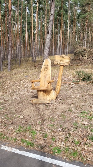Farbfoto: An einem Radweg am Waldrand steht eine Art Thron aus Baumstammteilen, in der Rückenlehne ist der Name Dieter eingebrannt und darunter die Zahl 87 in einem Lorbeerkranz. Daneben ist ein Schild an einem Ast des Baumstamms angebracht, darauf steht: "2023, [Fahrradsymbol], 2700 km". 