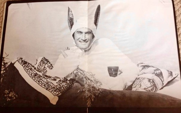 Man in a bunny suit lying on a pile of pillows holding a carrot in one hand and a cup of coffee in the other.
